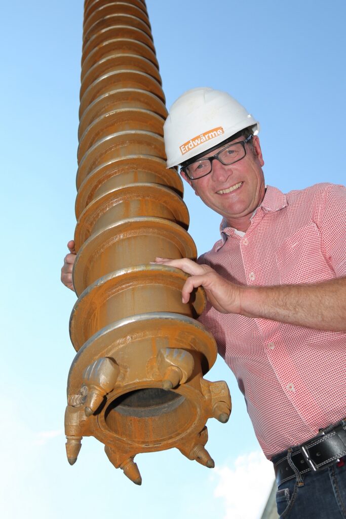 Ein Mann mit Schutzhelm und Brille steht lächelnd neben einem großen, rostfarbenen Industriebohrer, der für Erdwärmebohrungen verwendet wird, und hält ihn mit einer Hand fest. Im Hintergrund ist der blaue Himmel zu sehen.