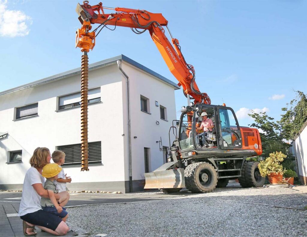 An einem sonnigen Tag beobachten eine Frau und ein Kind ein Baufahrzeug mit einem großen Bohraufsatz, das von einem Arbeiter in der Nähe eines modernen weißen Gebäudes bedient wird.