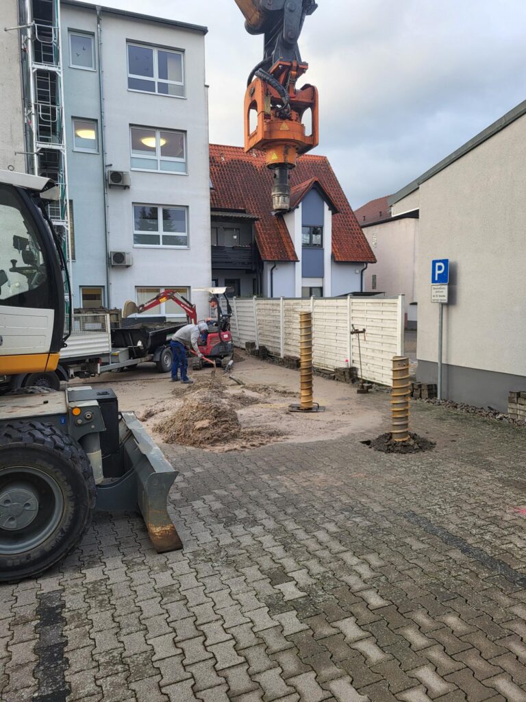 Baustelle mit Arbeitern, die Maschinen bedienen und Sand umschlagen. Zu sehen sind eine Bohranlage und Baumaschinen sowie zwei in den Boden eingebrachte Schraubpfähle. Im Hintergrund sind Mehrfamilienhäuser und ein Parkschild zu erkennen.