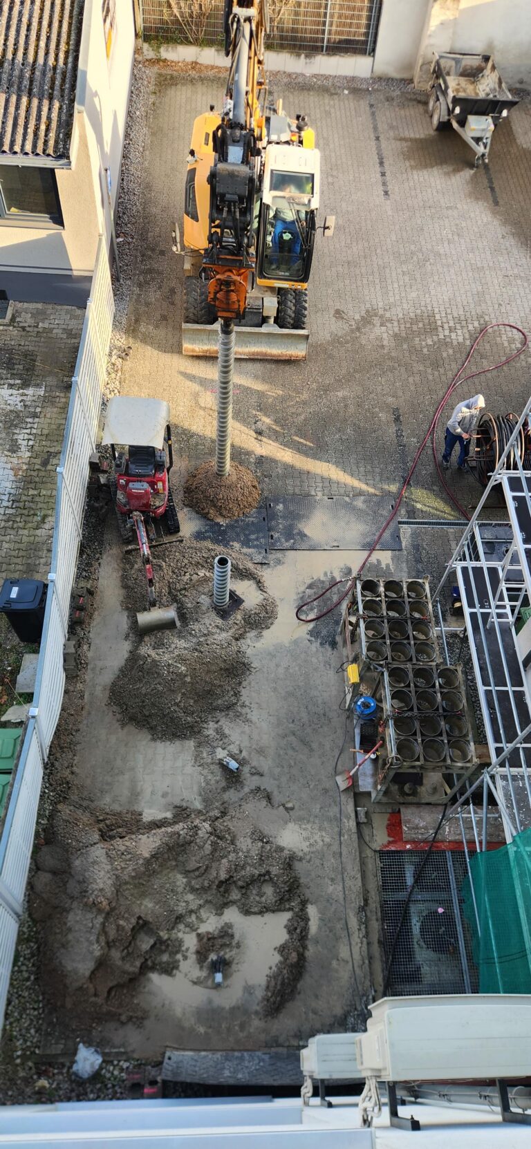 Eine Baustelle, auf der eine große Maschine in den Boden bohrt, daneben ein kleinerer roter Bagger, Rohre, Schlamm und Arbeiter, die mit den Maschinen hantieren – aus der Vogelperspektive betrachtet.