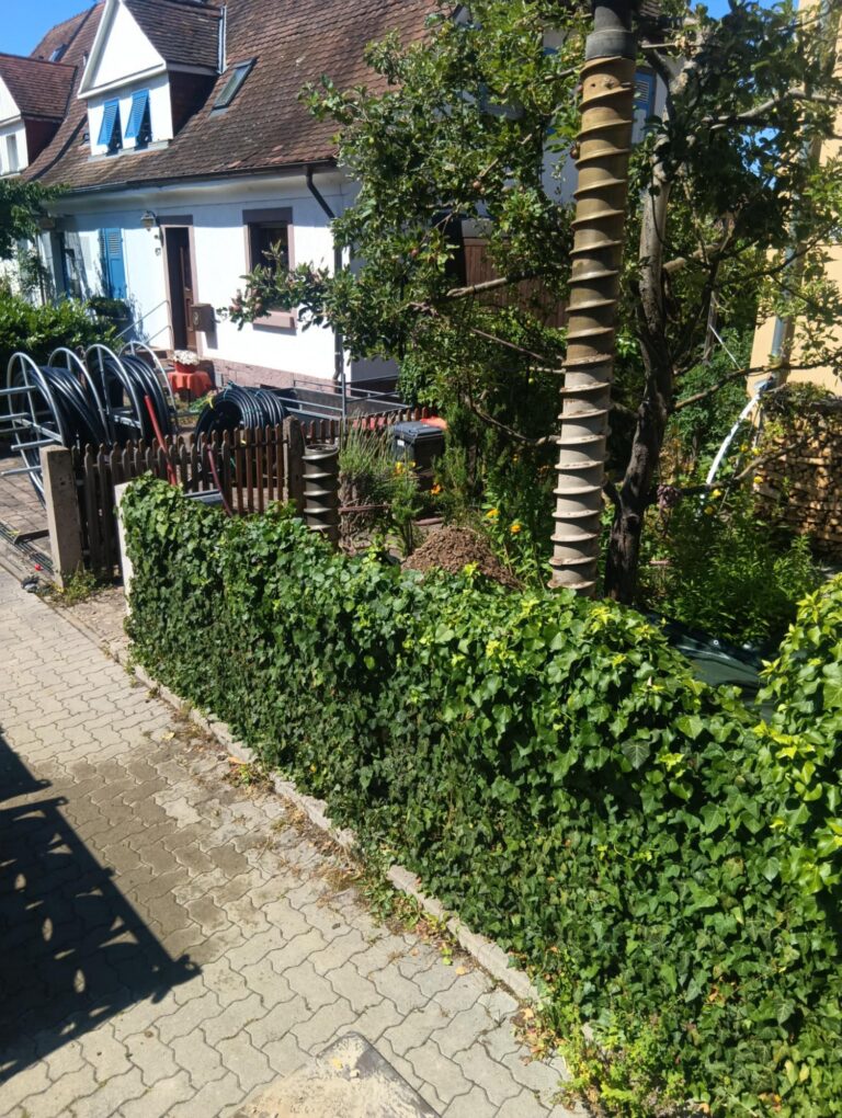 Ein sonniger Garten mit einer grünen Hecke, einem gepflasterten Weg und einem weißen Haus im Hintergrund. An einem hohen Baum sind am Stamm übereinanderliegende Scheiben zu sehen, und in der Nähe eines Holzzauns sind schwarze Rohre aufgerollt.