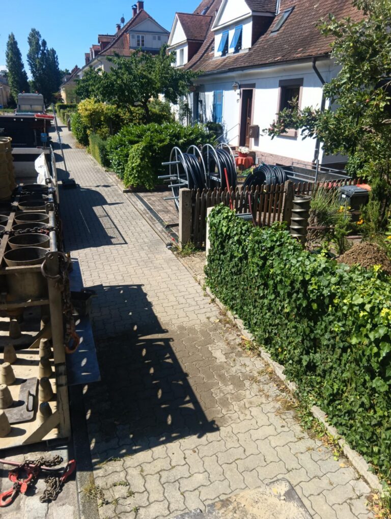 Eine Wohnstraße mit weißen Häusern, grünen Büschen und aufgerollten Kabeln in der Nähe eines Zauns. Auf dem gepflasterten Gehweg sind Schatten von Bäumen und Geräten zu sehen. Auf der linken Seite sind Baumaterialien zu erkennen.