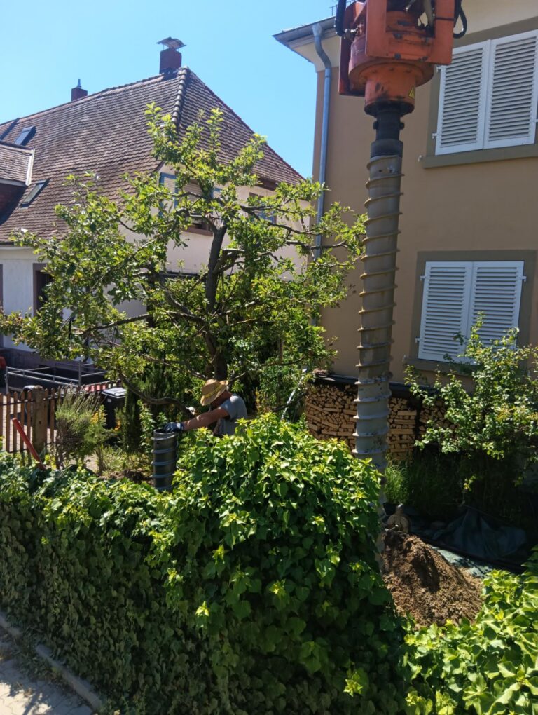 Eine Person mit Hut bedient an einem sonnigen Tag eine Bohranlage in einem Garten in der Nähe eines Hauses, dessen Zaun mit grünem Efeu bewachsen ist und neben dem ein Baum steht.