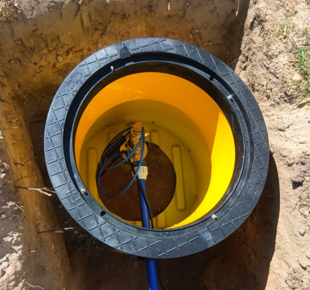 Eine Nahaufnahme eines gelben unterirdischen Tanks oder Brunnens mit schwarzem Rand, blauen Kabeln und Rohren im Inneren – möglicherweise Teil einer Grundwasserwärmepumpenanlage –, umgeben von ausgehobener Erde.