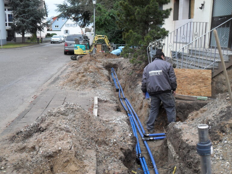Ein Arbeiter steht in einem Graben an einer Wohnstraße und verlegt blaue Versorgungsrohre. In der Nähe sind Baumaschinen und Baumaterialien zu sehen, am Gehweg liegen Erdhaufen, und im Hintergrund ist ein Haus mit einer Metalltreppe zu erkennen.
