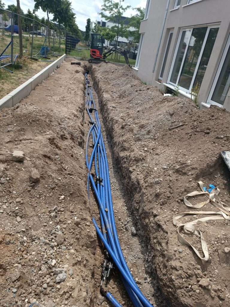 Mehrere blaue Rohre verlaufen durch einen langen, schmalen Graben, der neben einem Gebäude ausgehoben wurde; rund um die Baustelle sind Baumaschinen und loser Boden zu sehen.