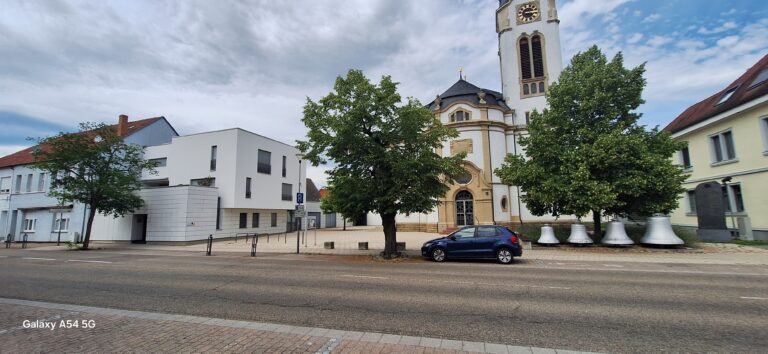 Ein blaues Auto steht auf einer Straße vor einer Kirche mit einem hohen Glockenturm. Neben dem Gebäude sind große Glocken zu sehen, und unter einem teilweise bewölkten Himmel säumen Bäume den Bürgersteig.