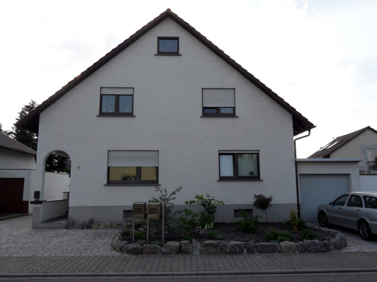 Ein graues, zweistöckiges Haus mit einem dreieckigen Dach, geschlossenen Fensterläden, einem kleinen Garten mit Steinen und Pflanzen davor, einer Einfahrt und einem silbernen Auto, das neben der Garage geparkt ist.