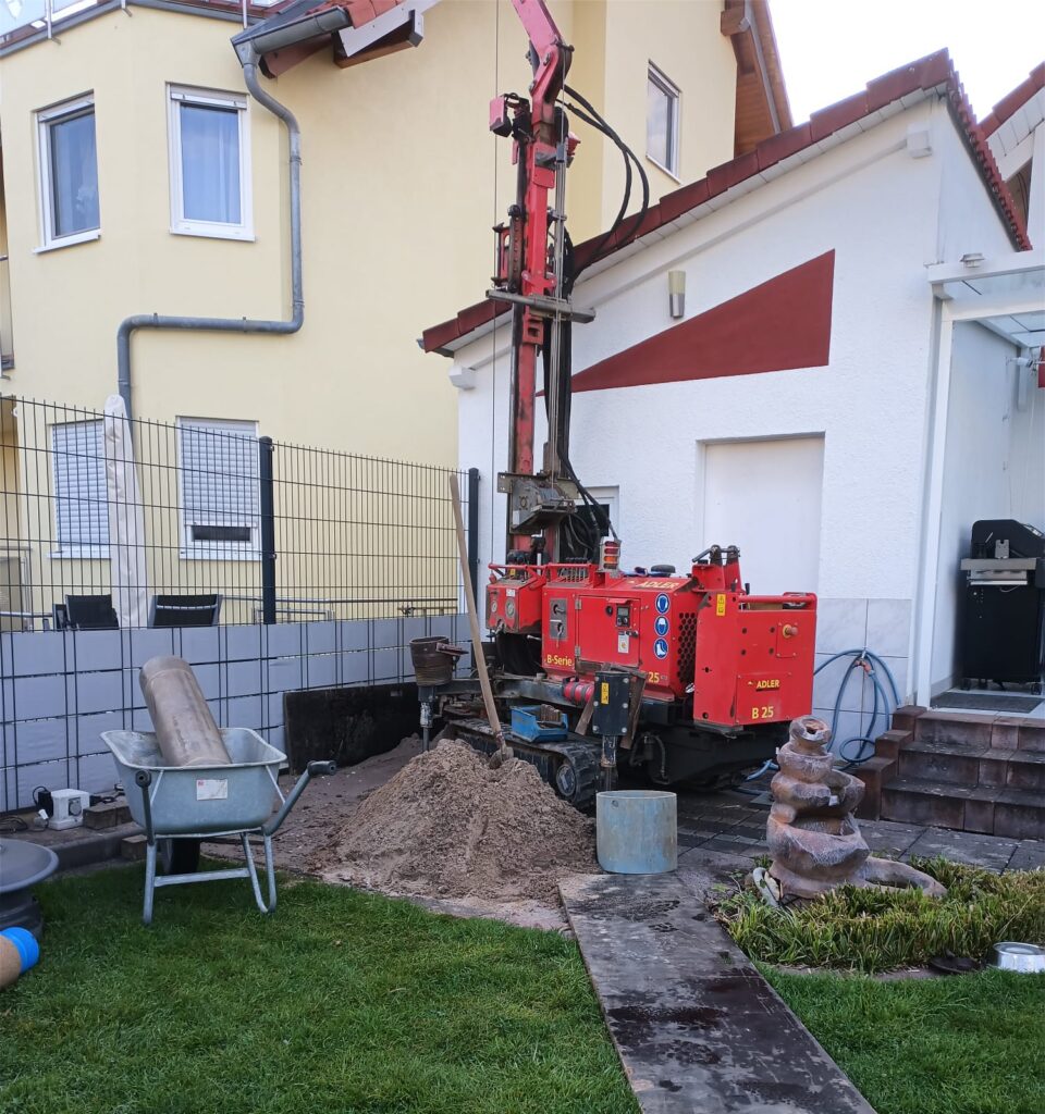 Neben einem Haus steht eine rote Bohrmaschine, umgeben von Sand, einer Schubkarre und Baumaterialien, was darauf hindeutet, dass im Garten gerade Bohr- oder Bauarbeiten stattfinden – möglicherweise für die Installation einer Grundwasserwärmepumpe.