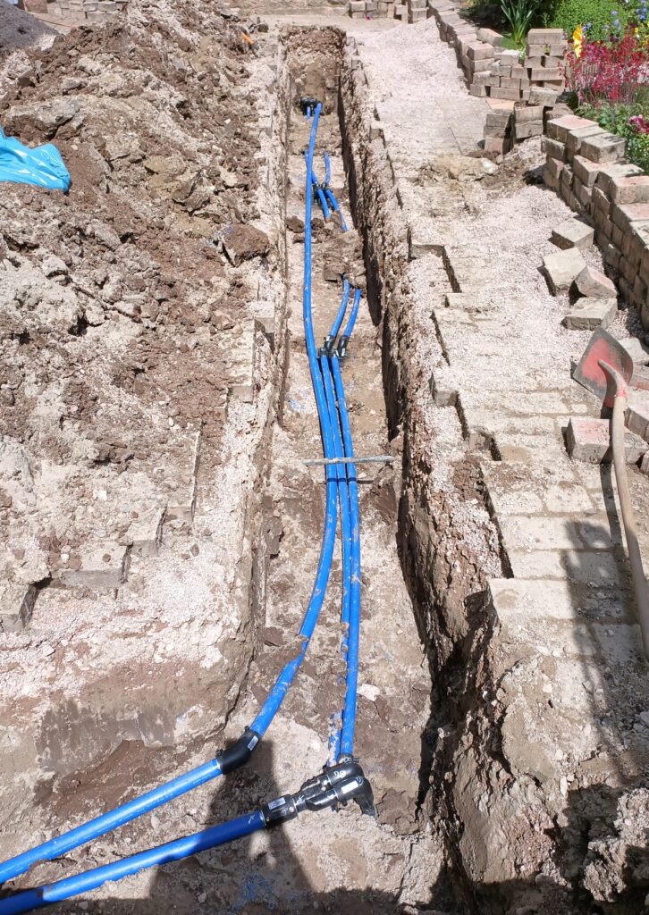 Ein Graben, in dem blaue Wasserleitungen und Erdwärmesonden verlegt sind, umgeben von Erdhaufen und Ziegelpflaster auf beiden Seiten. Im Hintergrund sind Gartengeräte und Pflanzen zu sehen.