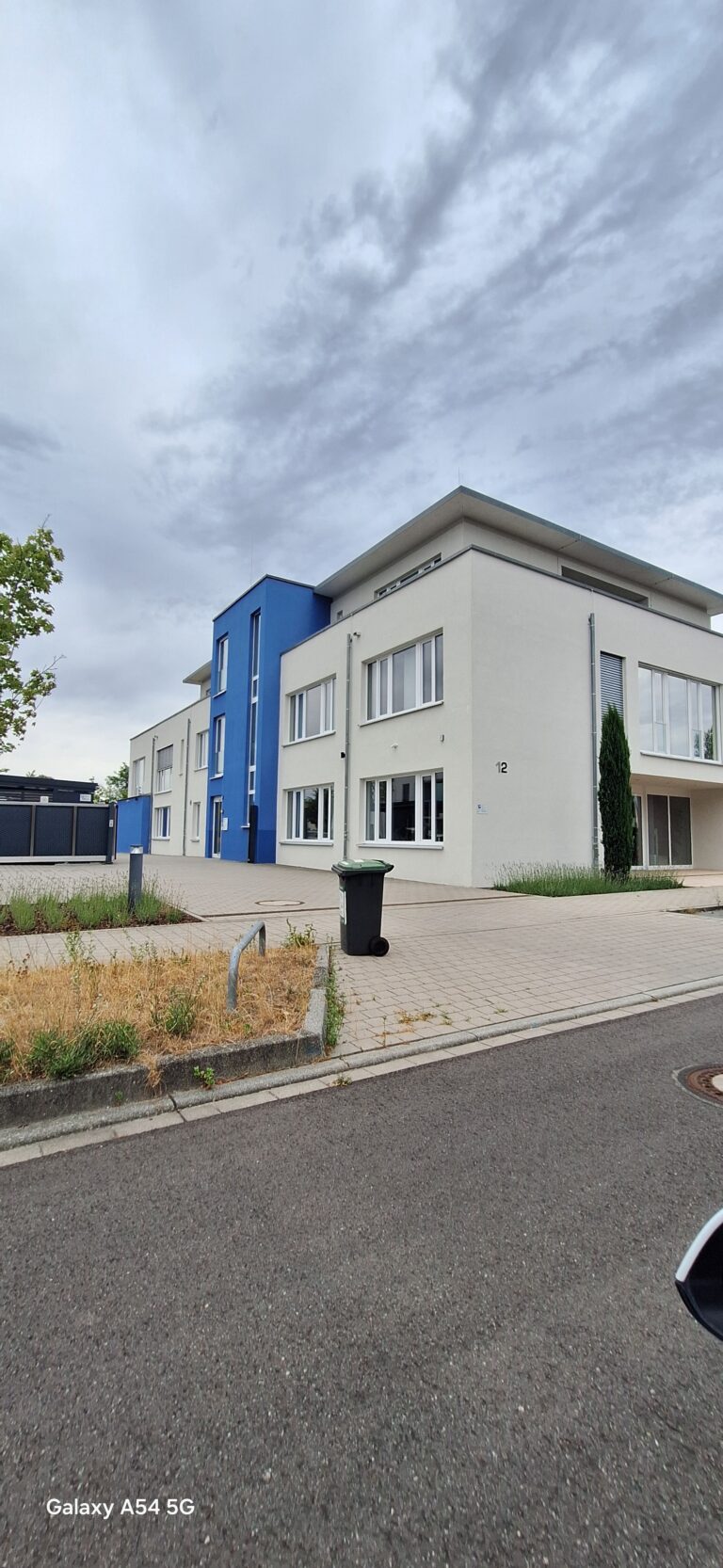 Modernes weiß-blaues Mehrfamilienhaus mit großen Fenstern, einem Flachdach und einer schwarzen Mülltonne auf der gepflasterten Einfahrt. Der Himmel ist bewölkt, und das Foto wurde von der Straße aus aufgenommen.