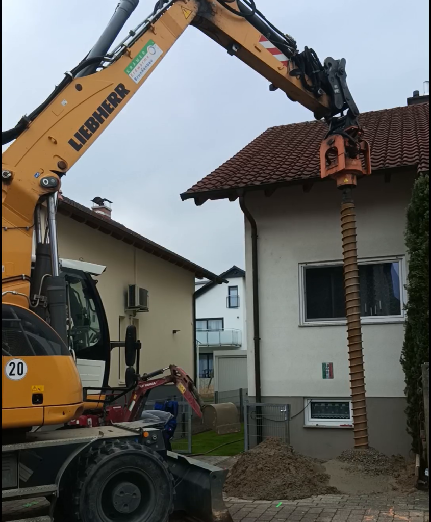 Ein gelber Bagger bohrt in der Nähe eines Hauses in den Boden, wobei sich sein großer Bohrstrang dicht am Ziegeldach befindet, um die Verlegung von Erdwärmesonden vorzubereiten. Am Fuß des Baggerarms türmt sich Erde auf, und im Hintergrund sind benachbarte Häuser zu sehen.