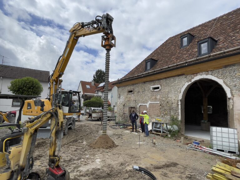 Eine Baustelle mit einem gelben Bagger, der in den Boden gräbt, drei Arbeitern in der Nähe und einem alten Steingebäude mit Ziegeldach im Hintergrund unter einem teilweise bewölkten Himmel.